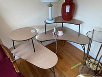 Two beige laminate mid-century modern tables, one corner triangular top, one elongated oval top, both with black legs, visible scratches and wear including burn mark and veneer chips.