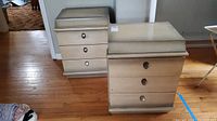 Two vintage wood nightstands shown side by side on hardwood floor, light beige finish with three drawers each, wood knob handles, showing wear including stains and scratches.