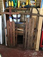 Photo of two vintage wooden sleds with metal runners and curved handle bars standing upright with rust and weathering.
