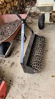 Full view of the tiller attachment with metal spiked roller elevating off the ground, showing handle and frame.