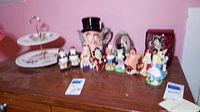 Wide view of Alice in Wonderland collectible figurines and decorative items on a wooden surface including salt and pepper shakers and a tiered dessert dish on the left.