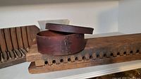 Photo showing two long wooden shaping molds with rounded cavities and a covered round wooden box with a lid on a white shelf.