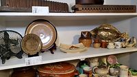 Shelf showing multiple pottery items including plates, pitchers, bowls, jugs, and decorative items arranged on two white shelves