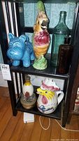 Shelf display showing the six items: blue ceramic dog, colorful Majolica parrot pitcher, brown and green glass bottles, Italian rooster pitcher, and other ceramics