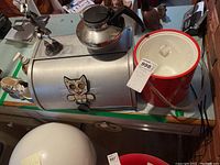 View showing metal breadbox with cat decoration, stainless steel coffee carafe, and red and white insulated ice bucket on countertop.