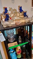 Multiple vintage glass bottles displayed on wooden racks and shelves including blue smiling man-shaped bottles and dog-shaped clear glass bottles.