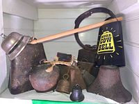 Overview of lot showing multiple iron and metal cowbells in various shapes and sizes, including one black bell with yellow text and a large rusted rectangular bell.