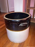 Full side view of vintage two-tone stoneware crock showing dark brown upper section and cream lower section on hardwood floor.
