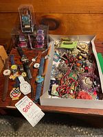 Photo shows multiple novelty watches and a box with assorted costume jewelry on a wooden table.