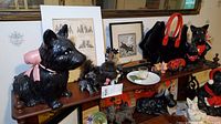 Photo showing the large black Scottie dog figurine with pink ribbon, smaller Scottie dog figurines, framed prints, Scottie dog handbag with red handles, small pigs figurines, and a dish with candle and stirrers on wooden shelf.