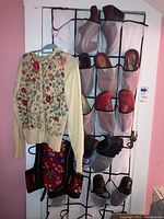 Vintage cream cardigan sweater with multi-colored floral embroidery hanging alongside a vintage red Yugoslavian embroidered midriff vest; over-the-door shoe organizer filled with various shoes including brown boots, red sandals, and slippers.