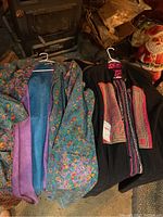 Two vintage jackets hanging on white hangers side by side, left jacket colorful floral pattern with blue and purple lining, right jacket black with embroidered front panel.
