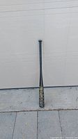 Full view of the black Easton Hammer baseball bat standing upright against a wall on pavement.
