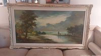 Front view of large, horizontal oil painting in ornate cream-colored frame depicting a landscape scene with trees, river, and sky