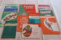 Photo of a collection of ten Christmas sheet music booklets and songbooks arranged on a white tablecloth.
