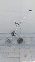 Photo of a lightweight manual push golf cart for golf bags, showing silver metal frame, two larger rear wheels and one smaller front wheel, and handle with grip and strap for securing golf bag.