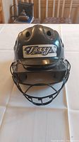 Front view of black youth helmet with attached metal face guard mask. 'Taye' branding visible on front.