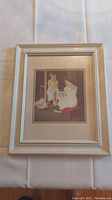 Framed Norman Rockwell print showing a woman in front of a mirror, framed in a light wooden frame with double matting.