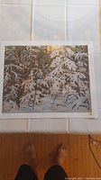 Photo showing the full art print of winter snowy woods on a white fabric surface with visible feet at the bottom of the frame.