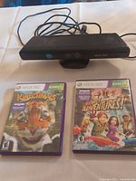 Black Xbox 360 Kinect sensor with cables and two Kinect game cases (Kinectimals and Kinect Adventures) on a white surface.