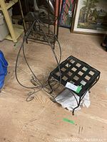 Two wrought iron plant stands on wooden floor: one with square glass top and lattice base, the other a hanging flower pot stand with curved legs