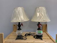 Two brass casting table lamps on wooden surface, both with plastic-covered off-white lampshades, cords visible and plugged in power source.