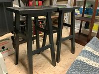 Two black wooden bar stools shown side by side on a wooden floor. Visible wear on edges and surfaces of tops and legs.