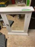Full front view of the white bathroom medicine cabinet with a rectangular mirror and metal latch on a wooden floor.