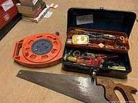 Black metal tool chest open showing assorted hand tools along with a large hand saw and an orange winding extension cord placed next to it