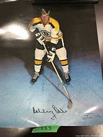 Full-length image of Bobby Orr in Boston Bruins uniform posed with hockey stick on ice, showcasing the autograph near the bottom of the poster.