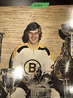 Color photo of Bobby Orr in Boston Bruins jersey posed with hockey trophies including the Stanley Cup.
