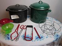 Two large enamel canning pots with lids in black speckled and green colors along with assorted red and green plastic and metal canning accessories including funnels, jar lifters, tongs, and a metal pot rack.