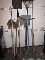 Overall lot image showing seven gardening tools propped against a garage door including two shovels, two rakes, garden hoe, garden fork, and small digging tool.