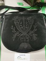 Front view showing black suede flap with floral embroidery and textured leather detail at bottom.