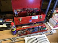 Open red metal Mastercraft tool box showing hand tools inside, a can of WD-40, and Stanley 100 Plus level on table.