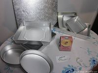 Nine-piece set of baking pans including square and round cake pans, two cake boards, and a boxed heating core accessory displayed on a table with patterned cloth.