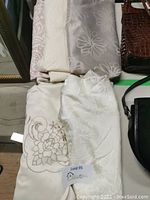 Lot of five folded assorted tablecloths showing variety in colors and patterns (cream, white, silver/taupe). Tablecloths stacked and visible fabric textures and designs.