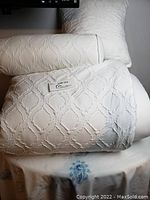 White queen size bedding displayed including pillows and bolster cushions with a geometric raised pattern.