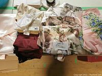 Photo showing folded clothing items including cream satin blouse, maroon skirt/pants with lace, and multiple floral print garments.