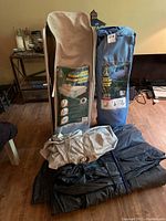 Two Woods folding beds in their storage bags, one beige (queen) and one blue (twin), placed on top of black and beige folded fabric pieces in a room with wooden floor and assorted furniture in background.
