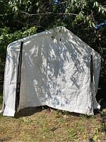 Front view of the white vinyl car port showing the entrance flap and overall structure under trees.