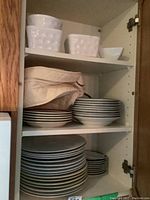Shelves with stacked white ceramic dinner plates, bread and butter plates, other small plates, and bowls. Includes a beige cloth bag and an additional white bowl on top shelf.