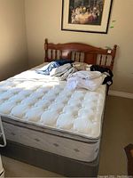 Full view of white quilted mattress, gray box spring and wooden headboard with linens piled on top