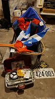Photo showing a lidded tote containing a blue and orange life jacket, two blue and white flags with fleur-de-lis design, wooden paddles with orange grips, a tackle box with fishing tackle, and two vintage license plates.