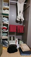 King size bedding folded and stacked on floor and shelves, showing variety of fabrics and colors including striped and floral duvet covers and pillow shams, assorted blankets and throws, towel stacks, and red curtain panels hung on rod.