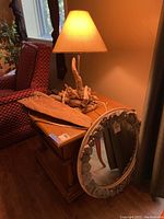Photo shows a wooden side table with a driftwood-based lamp with fabric shade lit. A rock-framed circular mirror leans against the table edge and a piece of driftwood plank sits flat on top.