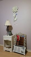 Wide view showing nightstand with lamp and urn on top, carved wood mirror on floor leaning on wall, small metal flower wall art hanging above nightstand, fan on floor, and wall background