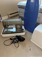 Full view of portable Singer sewing machine inside white plastic carry case, with black power cord and foot pedal, and a blue small shirt ironing board standing beside it.