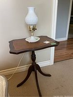 Side table with leather inlay top and white hobnail milk glass lamp on top near wall corner on carpeted floor.