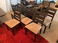 Six wood frame dining chairs with ladder backs and beige upholstery placed on a red area rug in a room with cardboard boxes and furniture.